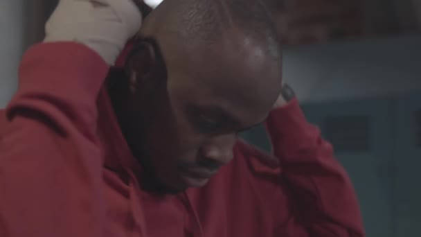 Κοντινό πλάνο τόξο πορτρέτο του 20-κάτι Αφρικής μαχητής των πολεμικών αθλημάτων με ξυρισμένο κεφάλι και τυλιγμένους καρπούς στέκεται στα αποδυτήρια, απογειώνεται κουκούλα του κόκκινο φούτερ του και κοιτάζοντας την κάμερα - Πλάνα, βίντεο
