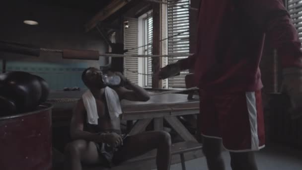Közepes lövés fiatal afro-amerikai bokszoló csupasz törzs ül padon mellett boksz gyűrű, edzőpartner besétál, leteszi kesztyűt, mindkét férfi ütő ököl, ivóvíz és csevegés - Felvétel, videó
