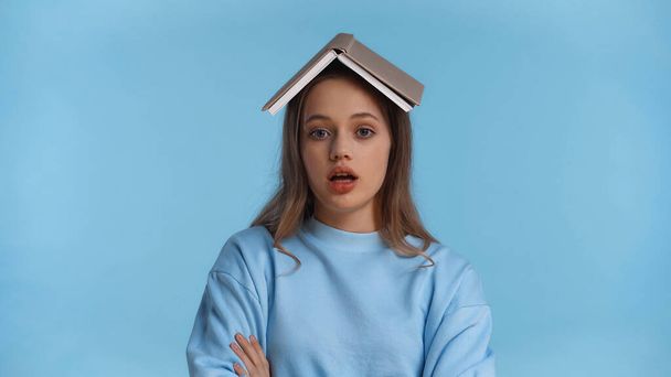 surprised teenage girl in soft sweatshirt with book on head isolated on blue - Photo, image