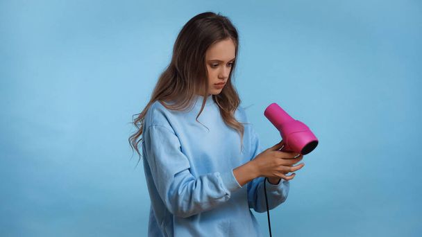 teenage girl in soft sweatshirt holding pink hair dryer isolated on blue - Фото, зображення