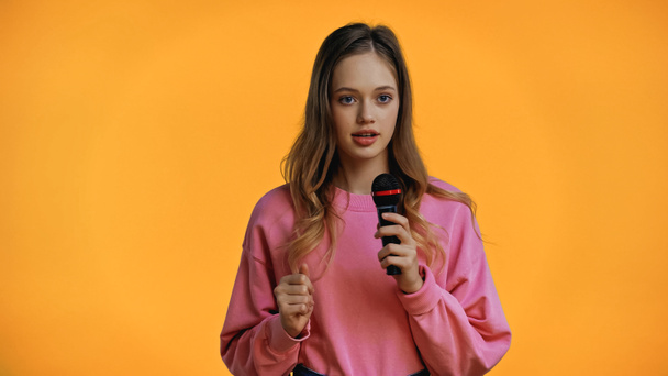teenage girl in pink sweatshirt talking while holding microphone isolated on yellow - Фото, изображение
