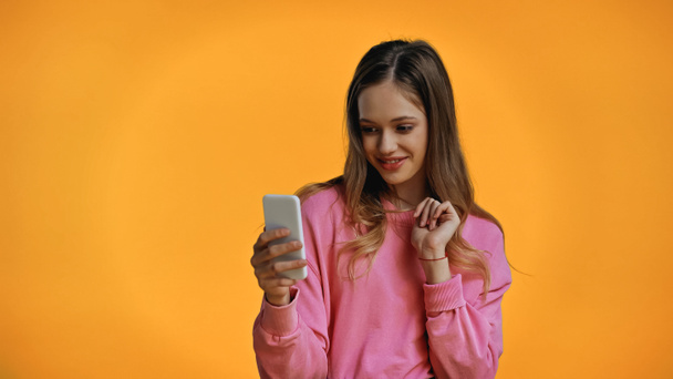 cheerful teenage girl in pink sweatshirt looking at smartphone isolated on yellow - Fotó, kép