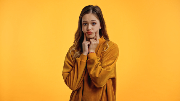 adolescente en suéter haciendo pucheros labios y tocando mejillas aisladas en amarillo - Foto, Imagen