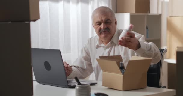 Ősz hajú férfi fehér ingben ellenőrzi a rendelést, amit egy vásárlónak küldenek laptoppal. férfi ül egy munkaasztalnál egy raktár háztartási vegyi anyagok. - Felvétel, videó