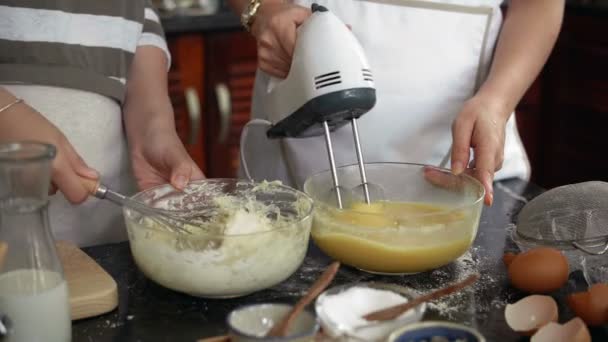 Αγνώριστη γυναίκα αναμιγνύει ζύμη μπισκότων με σύρμα ενώ ένα άλλο χτυπώντας τα αυγά με σύγχρονο μίξερ στην οικιακή κουζίνα - Πλάνα, βίντεο