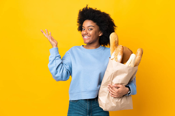 Joven mujer afroamericana comprando algo pan aislado sobre fondo amarillo extendiendo las manos a un lado para invitar a venir - Foto, Imagen