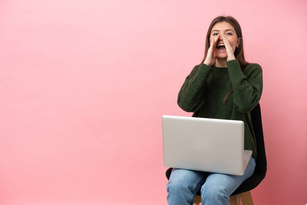 Genç beyaz kadın dizüstü bilgisayarıyla bir sandalyede oturuyor pembe arka planda izole edilmiş bağırıyor ve bir şey duyuruyor. - Fotoğraf, Görsel