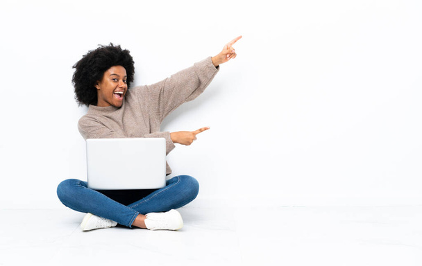 Jonge Afro-Amerikaanse vrouw met een laptop zittend op de vloer wijzend vinger naar de zijkant en presenterend een product - Foto, afbeelding