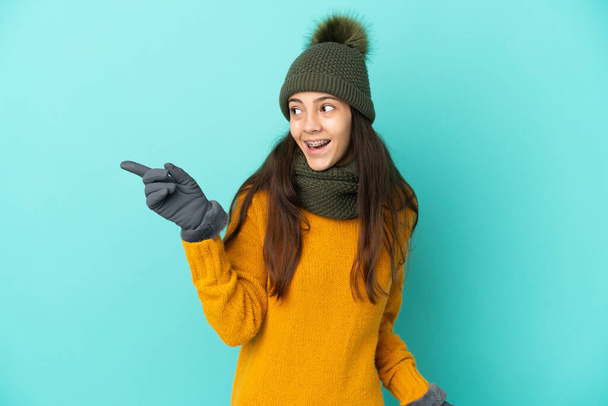 若いですフランス人の女の子は、指を持ち上げながら解決策を実現しようとして冬の帽子と青の背景に隔離されました - 写真・画像