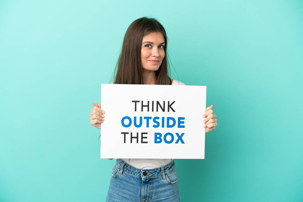 Молодая белая женщина, изолированная на синем фоне с плакатом с текстом "Думай вне коробки" - Фото, изображение