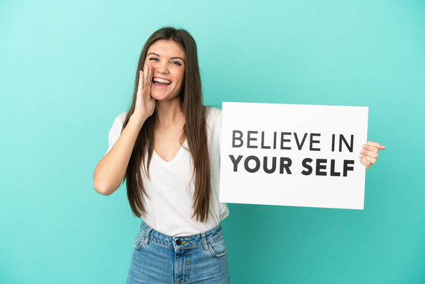 Young caucasian woman isolated on blue background holding a placard with text Believe In Your Self and shouting - 写真・画像