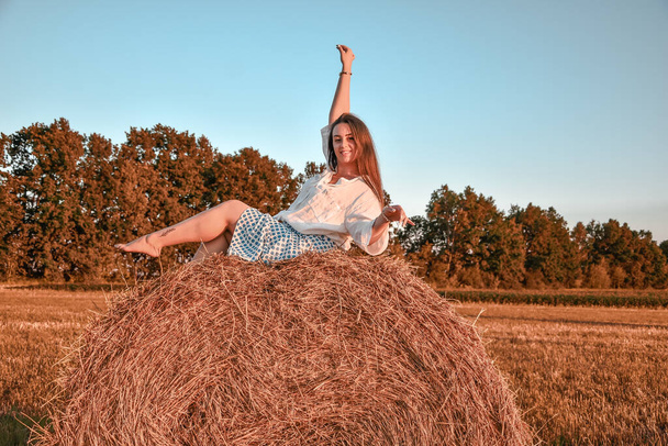 Portrait of young sexy woman on the haystack in morning sunlight, countryside. Beautiful woman in a dress sits on a haystack. Cottagecore - Photo, Image