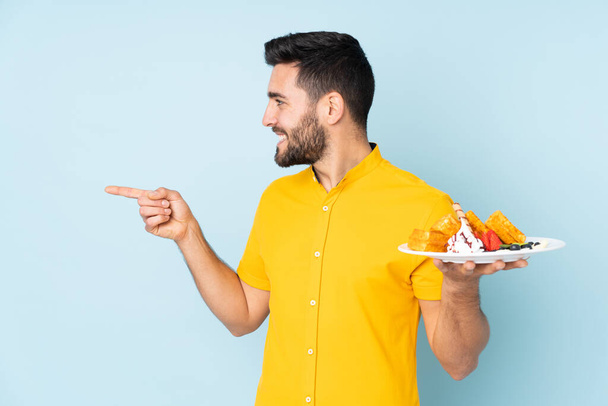 Caucasian man holding waffles isolated on blue background pointing to the side to present a product - Photo, Image