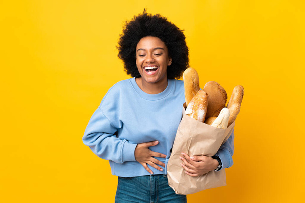 若いアフリカ系アメリカ人の女性が黄色の背景に隔離されたパンを買って多くの笑顔 - 写真・画像