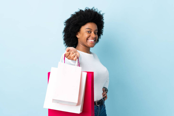 Young African American woman isolated on blue background holding shopping bags and giving them to someone - Photo, Image