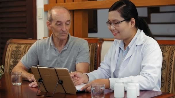Közepes lövés barátságos ázsiai orvos laboratóriumi kabát és szemüveg ül mellett érett férfi beteg, a tabletta számítógép és magyarázó recept - Felvétel, videó