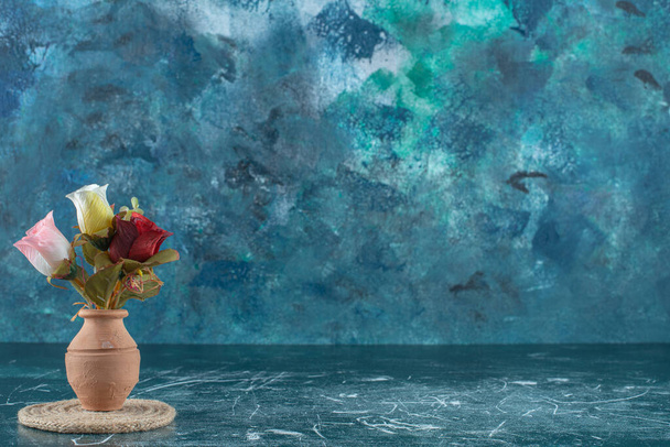 Künstliche bunte Rosen in einer Tonvase auf einem Untersetzer, auf blauem Hintergrund. Hochwertiges Foto - Foto, Bild