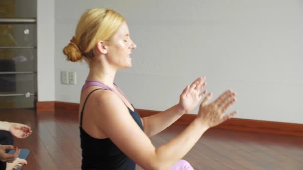 Blonde female teacher and group of women sitting on fitness mats in studio and practicing prayer pose - Footage, Video