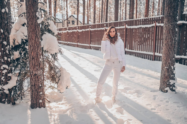 Zimní slunný studený portrét v sněžení krásné mladé ženy v bílém teplé oblečení dolů bunda na dvorku země dřevěný dům na pozadí zeleného smrku borovice pokryté sněhem. - Fotografie, Obrázek