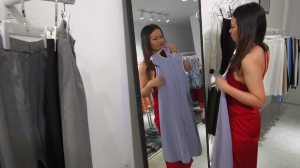 Média tiro de jovem mulher asiática em pé na frente do espelho, segurando vestido em suas mãos e olhando para si mesma com amplo sorriso - Filmagem, Vídeo