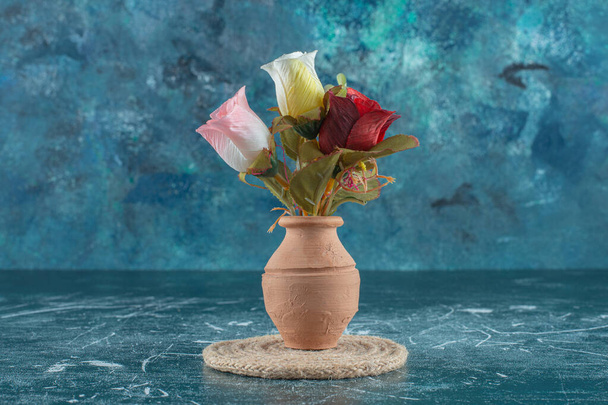 Artificial colorful roses in a clay vase on a trivet , on the blue background. High quality photo - Photo, Image