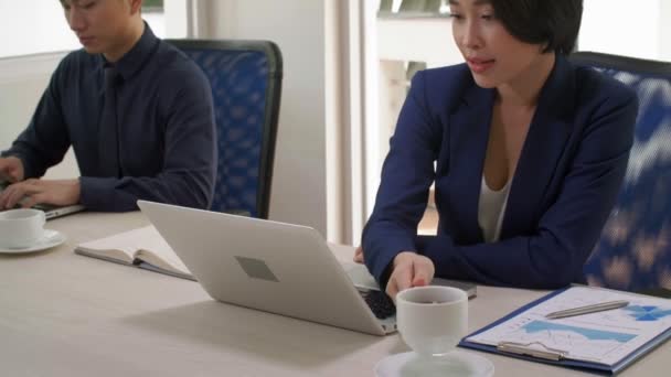 На передньому плані молода азійська бізнесменка сидить за робочим столом перед ноутбуком, п "є каву і бізнесмен на задньому плані працює з ноутбуком. - Кадри, відео