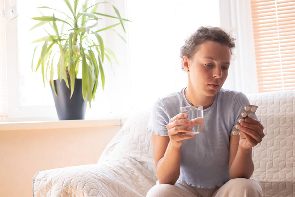 Θλιβερό κορίτσι που κρατά φάρμακα και ένα ποτήρι νερό σκέφτεται να πάρει φάρμακα, διαβάζοντας οδηγίες, δοσολογία του φαρμάκου, κάθεται στον καναπέ στο σπίτι. Αναρωτιέστε αν πρέπει να πάρετε αντικαταθλιπτικά χάπια - Φωτογραφία, εικόνα