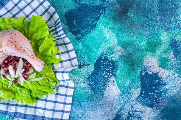Marinated chicken drumstick with pomegranate arils on a lettuce leaves on a board on a tea towel on the blue background. High quality photo - Photo, Image