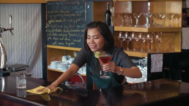 Middle-aged Asian female cafe owner cleaning bar counter with cloth thoroughly, serving cocktail Cosmopolitan and smiling at camera - Footage, Video