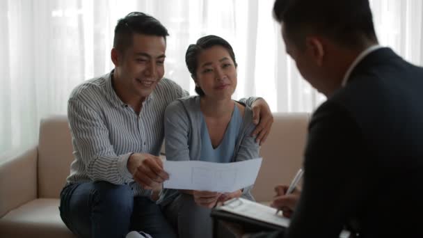 Junges asiatisches Ehepaar sitzt auf Couch und diskutiert Plan für neue Wohnung mit professionellem männlichen Immobilienberater - Filmmaterial, Video