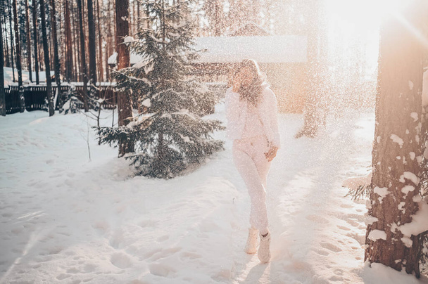 Зимовий сонячний холодний портрет в снігопаді красивої молодої жінки в білому теплому одязі в піджаку на задньому дворі заміського дерев'яного будинку на фоні зелених ялинових сосен, вкритих снігом
. - Фото, зображення