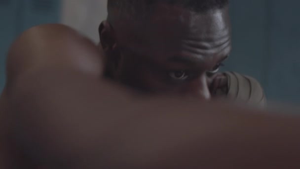 Primo piano di giovane pugile afroamericano con petto nudo e mano avvolge su polsi ombra boxe da solo in spogliatoio, praticare ganci, jab e uppercuts poi guardando la fotocamera - Filmati, video