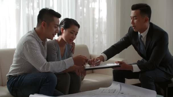 Young married Asian couple signing agreement with male real estate consultant and smiling happily while buying new apartment - Footage, Video