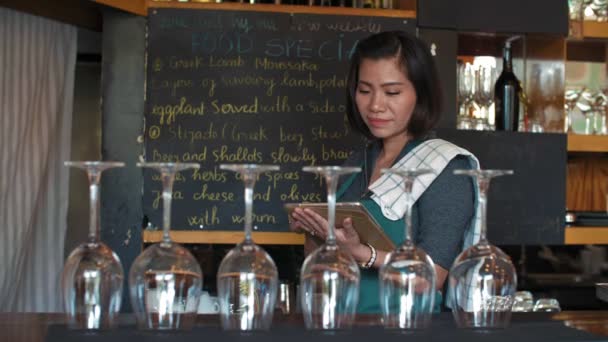 Attractive Asian female cafe owner standing at the bar, using digital tablet and smiling at camera happily, wine glasses placed in the foreground - Footage, Video