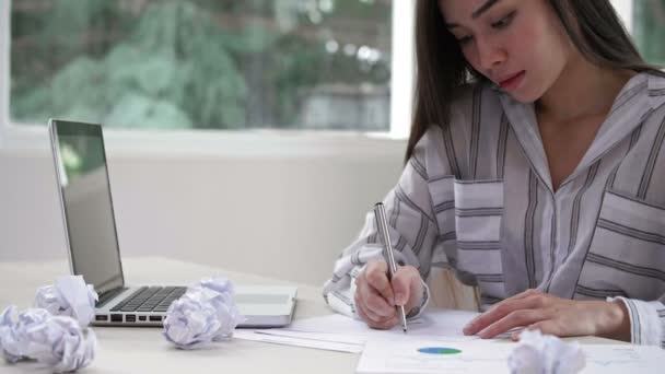 Közepes mennyiségű fiatal nő ül az asztalnál, tollat tart az egyik kezében, több levelet ír, majd áthúzza őket, és összegyűri a papírokat. - Felvétel, videó