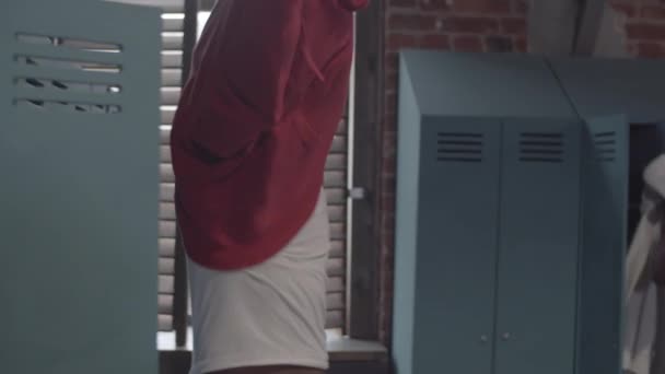 Schnappschuss eines fitten jungen Afroamerikaners mit rasiertem Kopf, in weißer Weste, der in der Umkleidekabine eines Fitnessstudios sein Sweatshirt auszieht und es in den Spind legt, während er mit einem Freund plaudert - Filmmaterial, Video
