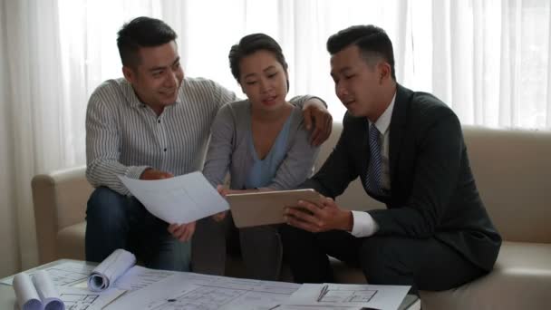 Consultor de propriedade masculina profissional mostrando plano de nova casa em tablet digital para jovem casal asiático sentado no sofá - Filmagem, Vídeo