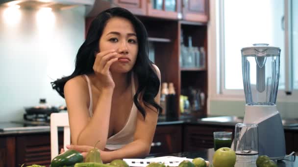 Elinde yazılı sözcüğü olan genç Asyalı kadın meyve suyu için meyve ve sebzelerle mutfak masasında oturmuş kameraya üzülerek bakıyor ve yardım ediyor.  - Video, Çekim