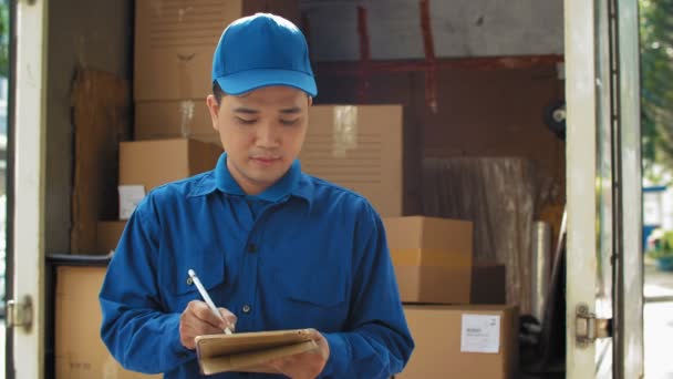 Średnie ujęcie azjatyckiego listonosza w niebieskim mundurze stojącego w pobliżu vana, piszącego tabletkę ze stylem, patrzącego w kamerę i uśmiechniętego - Materiał filmowy, wideo