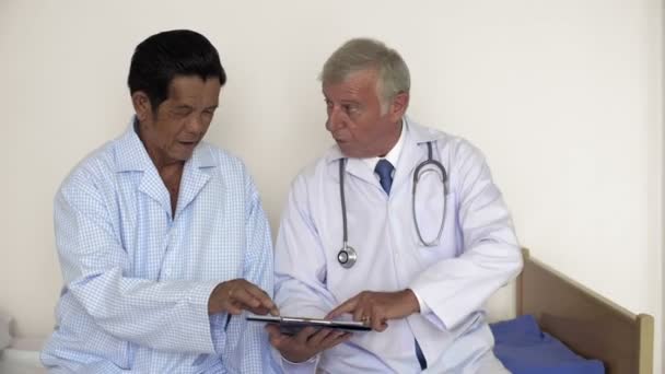 Anciano asiático con pijama consultando con un médico caucásico profesional sentado junto a él en la cama del hospital y explicando un tratamiento adicional  - Imágenes, Vídeo