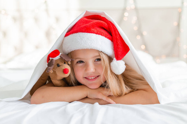 usmívající se holčička v Santa klobouku ležící na posteli s jelení hračkou - Fotografie, Obrázek