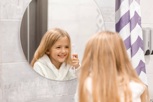 Χαριτωμένο κορίτσι με μπουρνούζι πλένει τα δόντια στο μπάνιο  - Φωτογραφία, εικόνα
