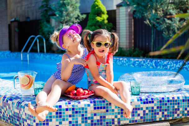 Cute girls sitting close to pool, having fun, eating fruits and drinking lemonade. Summer vacation concept  - Фото, изображение