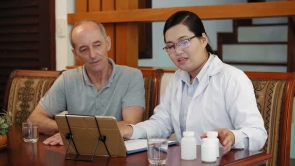 Colpo medio di amichevole medico femminile asiatico in camice da laboratorio e occhiali che mostrano bottiglie di pillole per maturare il paziente maschio e spiegare la prescrizione utilizzando tablet computer - Filmati, video