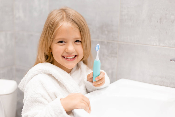 Χαριτωμένο κορίτσι με μπουρνούζι πλένει τα δόντια στο μπάνιο  - Φωτογραφία, εικόνα
