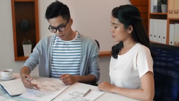 Young Asian male white collar worker discussing new project with young female colleague while sitting at office table and looking at business schemes - Footage, Video