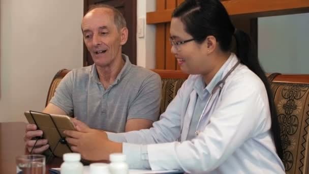Отслеживание среднего снимка оптимистической азиатки-врача в халате и очках, улыбающихся при разговоре с пациентом мужского пола, и использование планшетного компьютера для объяснения рецепта - Кадры, видео