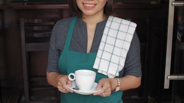Schnappschuss der attraktiven asiatischen Barkeeperin, die an der Bar steht, eine Tasse Kaffee in der Hand hält und fröhlich in die Kamera lächelt - Filmmaterial, Video