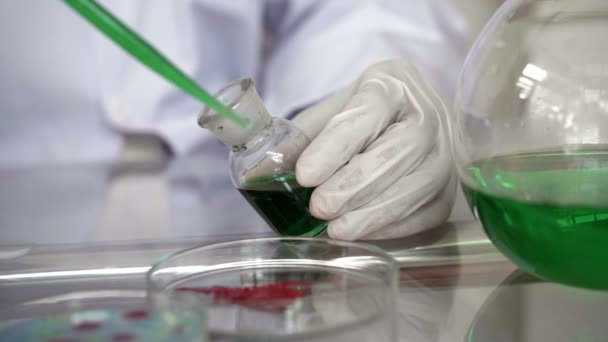 Primo piano delle mani in guanti con pipetta da laboratorio e liquido verde nella capsula di Petri riempita con soluzione media per un esperimento in un laboratorio di ricerca scientifica - Filmati, video
