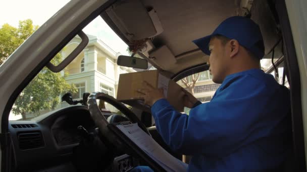 Κλείδωμα του ασιατικού courier σε μπλε στολή κάθεται στο αυτοκίνητο και γράφοντας πληροφορίες από δέμα σε έγγραφο - Πλάνα, βίντεο